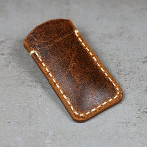 Puede incluir: Una funda de cuero marrón con costuras blancas. La funda tiene un acabado envejecido y una parte superior curvada. Las costuras son visibles alrededor de los bordes de la funda. La funda es rectangular y parece ser para un objeto pequeño.