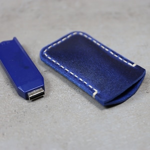 Puede incluir: Una funda de cuero azul con costuras blancas para un pequeño dispositivo electrónico. La funda está sobre una superficie gris.