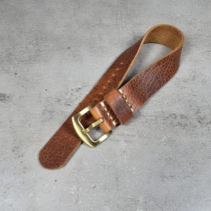 May include: A brown leather watch strap with a textured surface and a gold-colored buckle. The strap is stitched with white thread and has multiple holes for adjustment. The watch strap is shown on a gray surface.