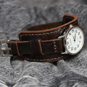 Op de afbeelding: Een bruin leren horlogebandje met een zilveren wijzerplaat waarop "CLASSIC" staat en zwarte cijfers. Het horloge ligt op een grijs stoffen oppervlak.