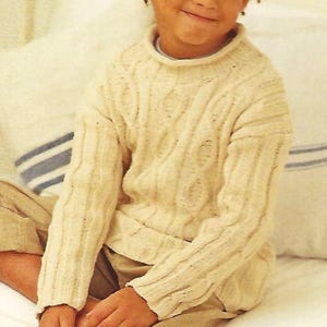 May include: A young person wearing a cream-colored cable-knit sweater and khaki pants sits on a white bed. The sweater has a ribbed collar and cuffs. The person is smiling and looking to the side.