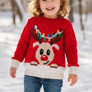 May include: A bright red long-sleeve knit sweater featuring a central graphic of a reindeer with a red nose and antlers decorated with colorful string lights. The sweater has a cream-colored ribbed hem and cuffs, paired with blue denim jeans.