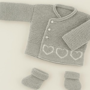 May include: A light gray knitted baby cardigan with three white heart designs on the bottom, and matching booties. The cardigan has three pearl buttons. The set is made of soft, textured yarn.