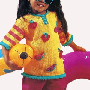 Może przedstawiać: Młoda osoba w żółtej dzianinowej koszulce z aplikacjami z czerwonych i zielonych owoców, w połączeniu z różowymi szortami. Koszulka ma paski na rękawach i turkusowe wykończenia. Akcesoria obejmują okulary przeciwsłoneczne, piłkę plażową i fioletowy nadmuchiwany koło.