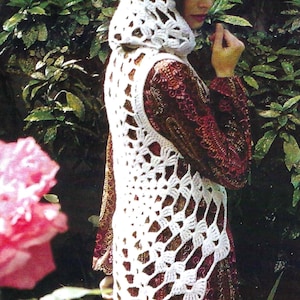 Könnte beinhalten: Eine weiße, offen gewebte Häkelweste und eine passende Kapuze. Die Weste hat ein geometrisches Muster und wird über einem gemusterten Kleid getragen. Der Hintergrund zeigt üppiges grünes Laub und eine rosa Rose.