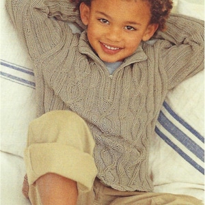 May include: A young person wearing a light brown cable-knit sweater with a zipper and matching khaki pants. The person is smiling and relaxing on a bed with a striped pillow.