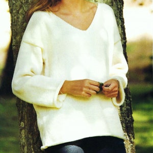 May include: A woman wearing a white knitted sweater with a v-neck and long sleeves. She is standing in front of a tree and has her hands clasped in front of her.