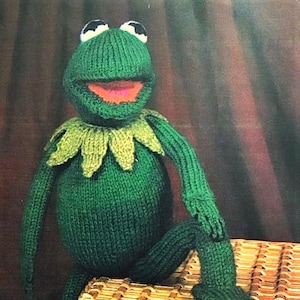 Puede incluir: Un títere de rana de punto verde con una gran sonrisa y ojos blancos. La rana está sentada en una cesta de mimbre.