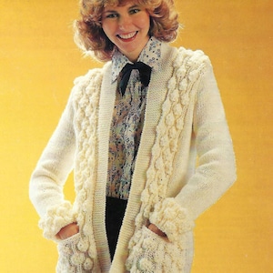 May include: A white knitted cardigan with a cable knit pattern and large, decorative buttons. The cardigan has pockets and a shawl collar.