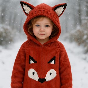 Könnte beinhalten: Ein Kind trägt einen rostfarbenen Strick-Fuchs-Hoodie mit Kapuze und Fuchsmotiv. Der Hoodie hat schwarze und weiße Akzente an Ohren und Gesicht. Das Kind trägt eine blaue Jeans und befindet sich in einer verschneiten Umgebung.