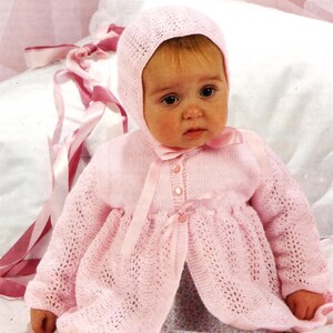 May include: A pink knitted baby outfit with a matching hat and booties. The outfit features a delicate lace pattern and a bow at the neckline.