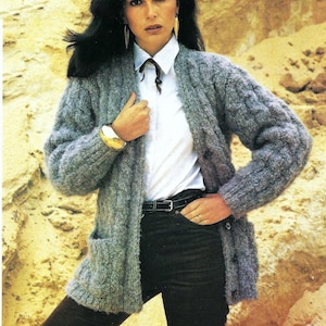 May include: A gray cable knit cardigan with a white collared shirt underneath. The cardigan has a button closure and two pockets. The model is wearing black pants.