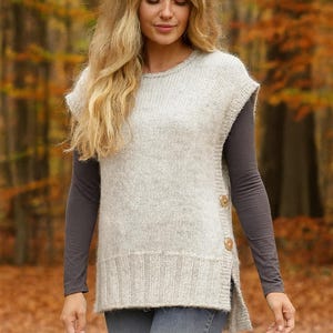 Peut inclure: Un gilet tricoté gris clair avec un col rond et des fentes latérales. Le gilet a des boutons en bois sur le côté et est porté sur une chemise grise à manches longues. L'arrière-plan est une forêt d'automne floue.