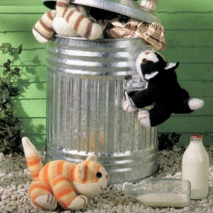 May include: A whimsical scene featuring knitted cat toys interacting with a metal trash can. One cat is inside, two are on top, and another is climbing the side. A bottle of milk and a tipped-over bottle are also present.