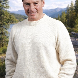May include: A cream-colored, crew-neck sweater. The sweater is made of a thick knit material. The person wearing the sweater is standing outside with a scenic background of a lake and mountains.