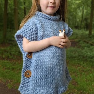 May include: A young child wearing a light blue knitted poncho with large wooden buttons down the side. The poncho has a high collar and short sleeves. The child is holding a small white figurine. The background is a blurred forest scene.
