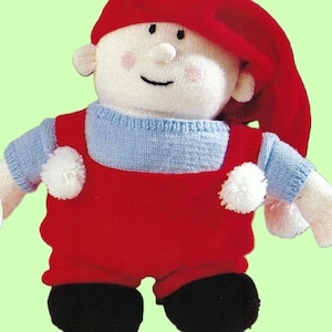 May include: A handmade knitted doll with a whimsical design. The doll features a red hat and overalls, a light blue sweater, and black shoes. White pom-poms adorn the overalls. The doll has a white face with rosy cheeks and a smiling expression.