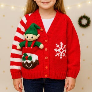 May include: A red knitted cardigan with a Christmas theme. The cardigan features a white snowflake design, a knitted elf in a pocket, and white and red striped sleeves. The cardigan has red buttons and is worn by a child.