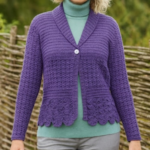 Puede incluir: Un cárdigan morado con un dobladillo festoneado y un cierre de un solo botón. El suéter de manga larga tiene un patrón de punto texturizado y un cuello chal. La persona de la imagen lleva un cuello alto turquesa y pantalones grises.