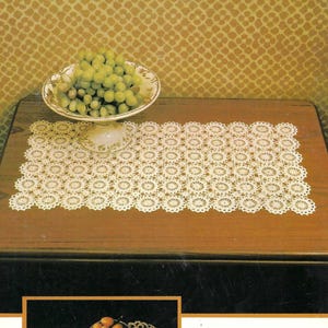 Puede incluir: Un camino de mesa de crochet blanco con un patrón circular repetitivo. El camino de mesa está sobre una mesa de madera con un cuenco de uvas verdes sobre un pedestal. La imagen es de un libro de patrones de crochet vintage.