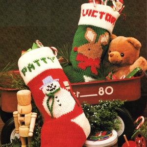 Puede incluir: Dos calcetines de Navidad tejidos con nombres personalizados, "VICTOR" y "MITTY", están llenos de bastones de caramelo y se exhiben en un vagón rojo con un cascanueces de madera y otras decoraciones navideñas.