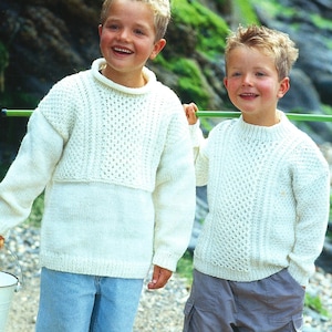 May include: Two young boys wearing white knitted sweaters with a cable knit pattern. The boy on the left is holding a white bucket. Both boys are wearing shorts and are barefoot.