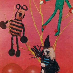 May include: A Halloween party craft pattern featuring three knitted toys: a bee, an elf, and a witch. The bee is black and yellow with pink stripes. The elf is green with red and white accents. The witch is black with a blue and white striped dress and a black hat.