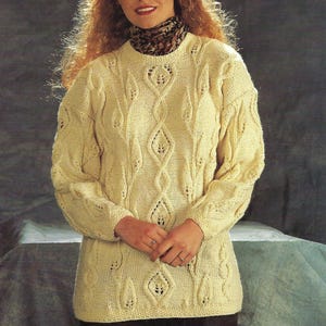 May include: Cream-colored, long-sleeved sweater with a cable-knit design featuring leaf-shaped patterns. The sweater has a crew neck and is worn over a patterned turtleneck. The overall style is classic and cozy.