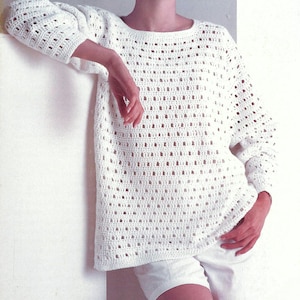 May include: A white, open-weave crochet sweater with long sleeves. The sweater has a loose, boxy fit and is paired with white shorts. The crochet pattern features rectangular openings throughout the design, creating a textured look.