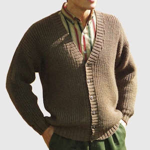 May include: A brown button-down cardigan with a ribbed texture. The sweater is worn over a striped shirt with red, green, and tan stripes. The person is wearing green trousers. The cardigan has a V-neck and long sleeves.