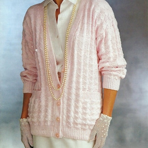 May include: A light pink cardigan with a textured knit pattern, worn over a white shirt and skirt. The cardigan has pockets and button closures. A pearl necklace and a white hat complete the ensemble. The gloves are white.