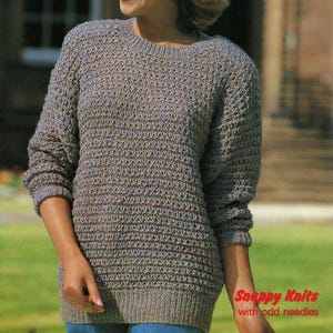 May include: A woman wearing a grey knitted jumper with a textured pattern. The jumper has long sleeves and a ribbed hem. The text "Snappy Knits with odd needles" is in the bottom right corner. The jumper is a neutral colour.