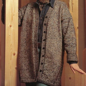 May include: A long, button-down cardigan in a speckled brown and beige knit. The cardigan is worn over a denim shirt and a patterned scarf. The model is standing next to a wooden wall.