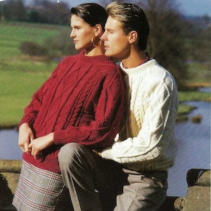 May include: A couple wearing matching cable knit sweaters. The woman is wearing a red sweater and a plaid skirt. The man is wearing a white sweater and gray pants. They are standing in front of a stone wall.