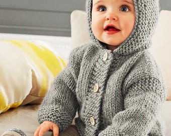 May include: A gray knitted hooded cardigan with button closures, worn by a baby sitting on a white bed.