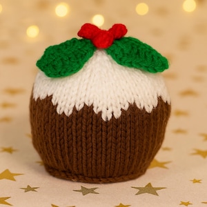 May include: A knitted Christmas pudding, featuring a brown base, white top, and green and red holly leaves. This handmade decoration is a festive holiday item, ideal for seasonal decor. The pudding is a soft, textured item.