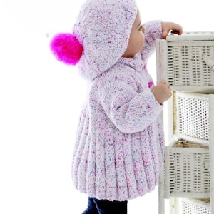 May include: A white knitted baby jacket with a pink pom-pom on the hood. The jacket has a speckled pattern and is worn by a baby standing next to a white wicker basket.