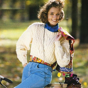 Könnte beinhalten: Eine Frau trägt einen weißen Strickpullover mit Zopfmuster, einen blauen Schal, einen bunten Gürtel und eine blaue Jeans. Sie hält einen bunten bestickten Schal mit Quasten.