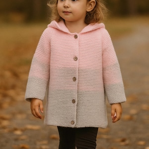 Può includere: Un cappotto con cappuccio e bottoni rosa e grigio. Il cappotto in maglia è lungo fino al ginocchio. La bambina indossa leggings scuri e stivali marroni.