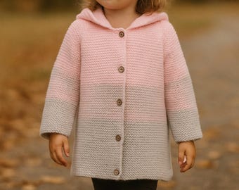May include: A hooded, button-down coat in a pink and gray color block design. The coat is knee-length and appears to be knitted. The child is wearing dark leggings and brown boots.