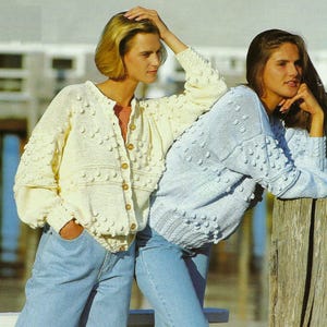 May include: Two women wearing knitted cardigans with a textured bobble design. One cardigan is white and the other is light blue. Both women are wearing blue jeans.