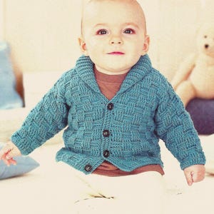 May include: A baby wearing a teal blue knitted cardigan with a textured weave pattern. The cardigan has a shawl collar and is buttoned with dark brown buttons. The baby is also wearing a brown shirt and white pants.