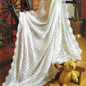 Puede incluir: Un chal blanco, intrincadamente tejido con un borde festoneado, está drapeado sobre un soporte de madera. El chal presenta un patrón calado detallado. Un oso de peluche vintage está sentado en un pequeño taburete de madera cercano.