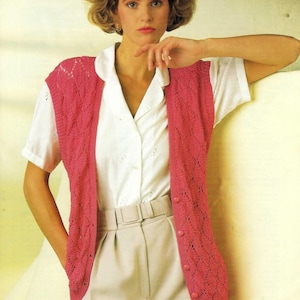 May include: A pink crocheted sleeveless vest with a diamond pattern and button closures. The vest is worn over a white button-down shirt and beige pants.