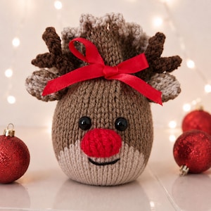 May include: A brown knitted reindeer ornament with a bright red nose, black button eyes, and dark brown antlers. The top is gathered with a vibrant red satin ribbon tied in a bow. The lower face is a lighter beige colour with a stitched black mouth.