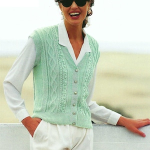 May include: A pale green knitted vest featuring a cable knit pattern, button fastening, and a V-neck. The vest is worn over a white collared shirt and paired with white trousers. The style is classic and preppy.