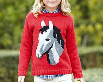 May include: A young person wearing a red knitted sweater with a white and gray horse design. The sweater has long sleeves and a crew neck. The person is holding a green apple in their hand.