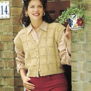 May include: A woman wearing a tan, button-up, knitted vest over a collared shirt and red pants. The vest has a square pattern. The woman is smiling and standing near a brick wall with a floral planter.