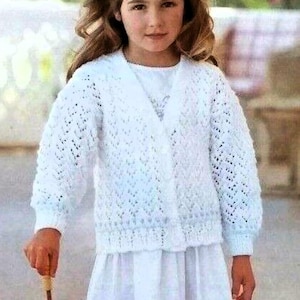 May include: A white knitted cardigan with a delicate openwork pattern. The cardigan has a V-neckline and button closure. The model is wearing a white dress and holding a white umbrella.