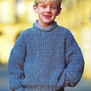 Puede incluir: Un niño pequeño con un suéter azul tejido con un patrón texturizado. Está sonriendo y tiene el pelo rubio.